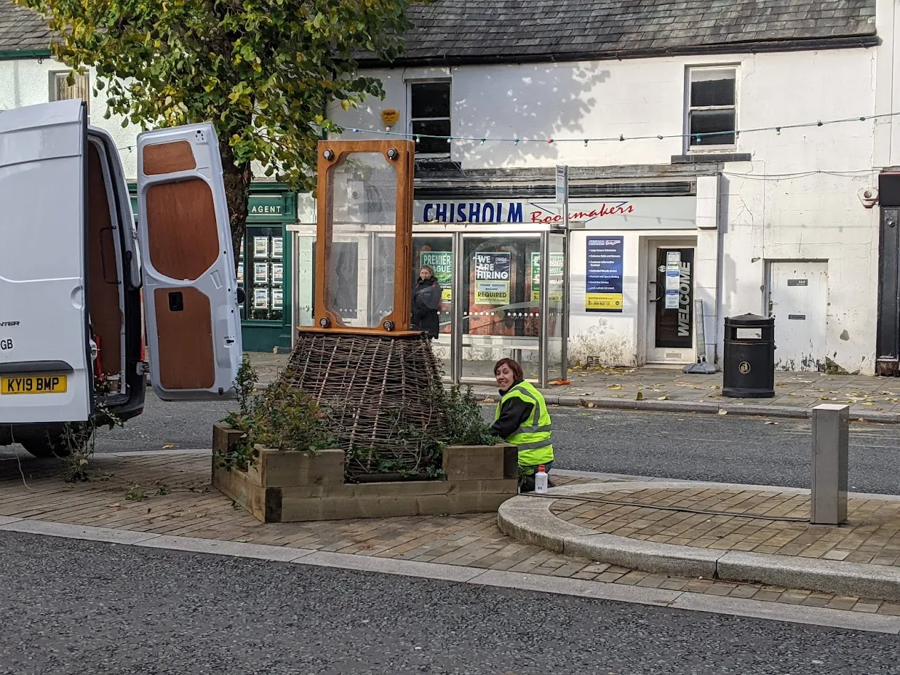 Woman kneeling on ground next to plants and wooden structure.