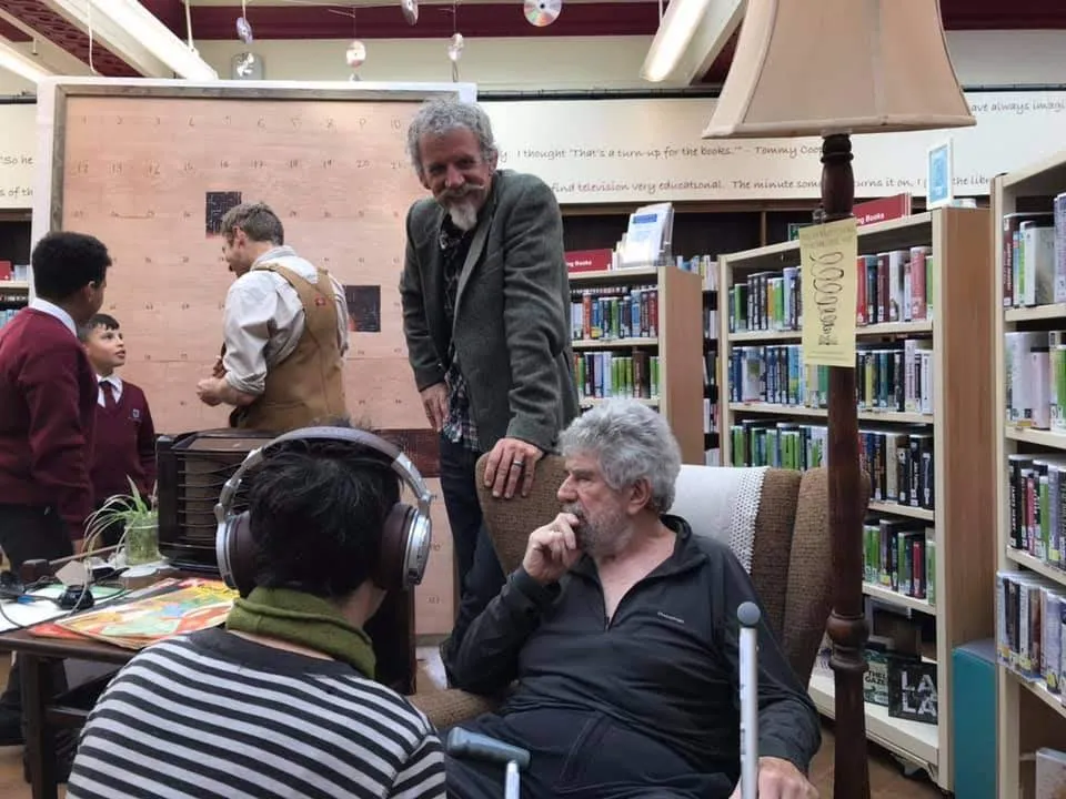 Person listening to headphones, man sitting, people in background.