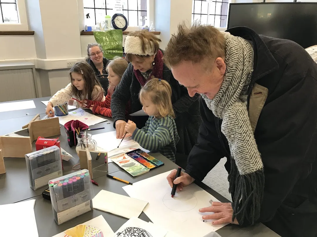 Group of grown-ups and children drawing and painting leaning over a table covered in sheets of paper, paints, pens and mirrors.
